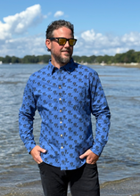 Load image into Gallery viewer, Man wearing a blue patterned shirt standing by a body of water with trees in the background.